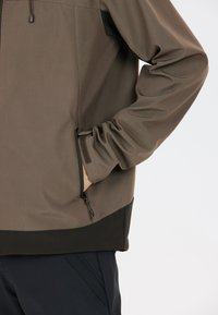 Braune Jacke mit glatter Oberfläche, ausgestattet mit einer Reißverschlusstasche, elastischen Bündchen und einem kontrastierenden schwarzen Saum am unteren Rand.
