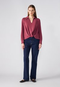 Langermet, V-hals maroon bluse med vridd front, paired med mørkeblå slengbukser. Modellen har på seg svarte flats.