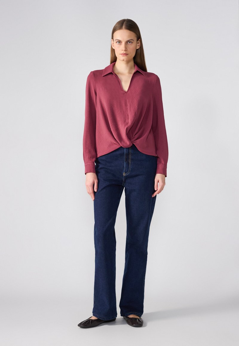 Bordowa bluzka z długim rękawem i dekoltem w kształcie litery V, z przekręconym przodem, zestawiona z ciemnoniebieskimi dzwonami. Modelka nosi czarne płaskie buty.