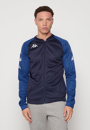 Giovane uomo che indossa una giacca sportiva con zip blu navy e blu con motivi di logo bianchi sulle maniche e pantaloni della tuta grigio chiaro, in piedi davanti a uno sfondo semplice.