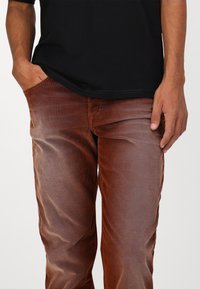 Barna bordás nadrág, slim fit fazonban, elülső zsebbel és látható varrással, fekete pólóval párosítva. Puha tapintás és finom fényesség jellemzi.