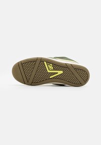 Semelle de sneaker verte et beige avec caoutchouc texturé pour une meilleure adhérence, présentant un motif circulaire et un accent de logo jaune près du talon.