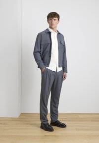 Gray tailored jacket with two chest pockets, worn over a white shirt, paired with gray pleated trousers and black shoes, standing on wooden floor.