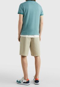 Osoba v tyrkysovém polo tričku, béžových šortkách a bílých teniskách s tyrkysovými, námořnickými a oranžovými akcenty, stojí na jednotném pozadí.