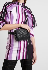 Czarna pikowana skórzana torba z wytłoczonym logo, noszona w talii. Modelka ma na sobie sukienkę w wielokolorowe paski z bufkami.