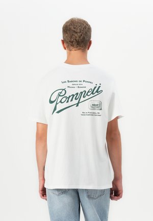 Young man with short curly hair wearing a white T-shirt with green Pompeii soap brand text and light blue jeans, facing away.