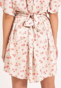 Kwiatowa sukienka w jasnym beżu z małymi różowymi i czerwonymi kwiatami. Posiada związaną kokardę z tyłu oraz zwiewną, sięgającą kolan spódnicę z miękkiego materiału.