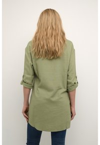 Zeleno laneno srajco z zvijanimi rokavi in daljšim zadnjim robom. Gladka tekstura in sproščen kroj, v kombinaciji z modrimi kavbojkami.