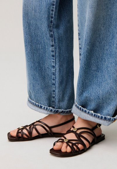 Piedi con sandali piatti marrone scuro con cinturini e dettagli dorati, abbinati a jeans azzurri chiari risvoltati su sfondo neutro.