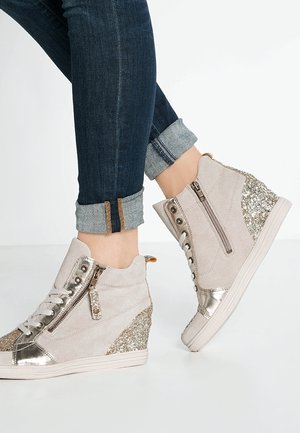 Füße in beigefarbenen Keil-Sneakern mit goldenen Glitzerakzenten, Reißverschlüssen und Schnürsenkeln, kombiniert mit hochgekrempelten dunkelblauen Jeans.