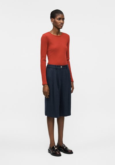 Γυναίκα στέκεται φορώντας εφαρμοστό μακρυμάνικο κόκκινο τοπ, μπλε σκουρόχρωμη πλισέ φούστα μέχρι το γόνατο και μαύρα μοκασίνια, με απλό φωτεινό φόντο.