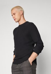 Musthave mustrilõngaga must kampsun, ümmarguse kaeluse ja ribapäistega, tekstuurse mustriga, koos hallide ruuduliste pükstega.