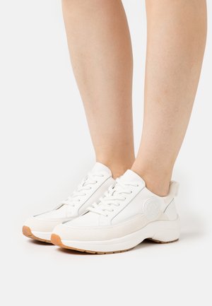 Benen met witte vetersneakers met beige accenten en een bruine zool op een witte achtergrond.