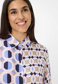 Donna sorridente che indossa una camicia a maniche lunghe con motivi geometrici in rosa pastello, blu, beige e nero, e piccoli orecchini a cerchio d'oro.
