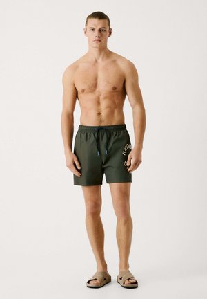 Pantalones de baño caquis con cintura elástica, cordón azul y detalle de logo blanco. Llevados con sandalias de tira beige. Se ve piel.