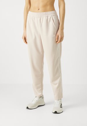 Žena nosící světle růžové tepláky Adidas s elastickým pasem a bílé tenisky Adidas, stojící na jednobarevném pozadí.