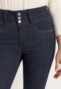 Korkeavyötäröiset tummansiniset denim-farkut, joissa on kolme metallipainiketta, hieman joustavaa materiaalia ja etutaskut. Sileä pinta ja siisti ompeluyksityiskohdat.