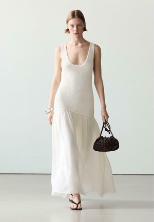 Frau trägt ärmelloses weißes Strickkleid mit fließendem Rock, hält schwarze gewebte Handtasche, trägt Armreifen und schwarze Sandalen, geht vorwärts.