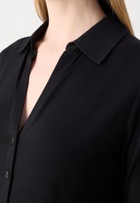 Gros plan d'une personne portant une chemise noire à col avec des boutons, avec de longs cheveux raides visibles sur un côté.