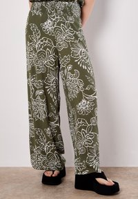 Pantaloni fluidi verde oliva con motivi floreali bianchi; vita smock; design a gamba larga; indossati con sandali platform neri.