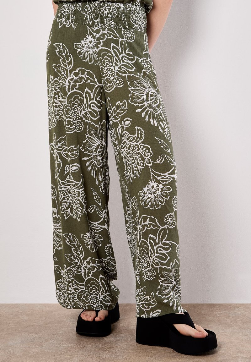 Pantaloni fluidi verde oliva con motivi floreali bianchi; vita smock; design a gamba larga; indossati con sandali platform neri.