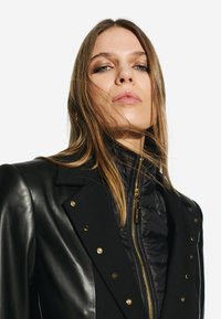 Chaqueta de cuero negro y tela acolchada con herrajes dorados, diseño entallado y parte delantera cruzada con broches visibles.