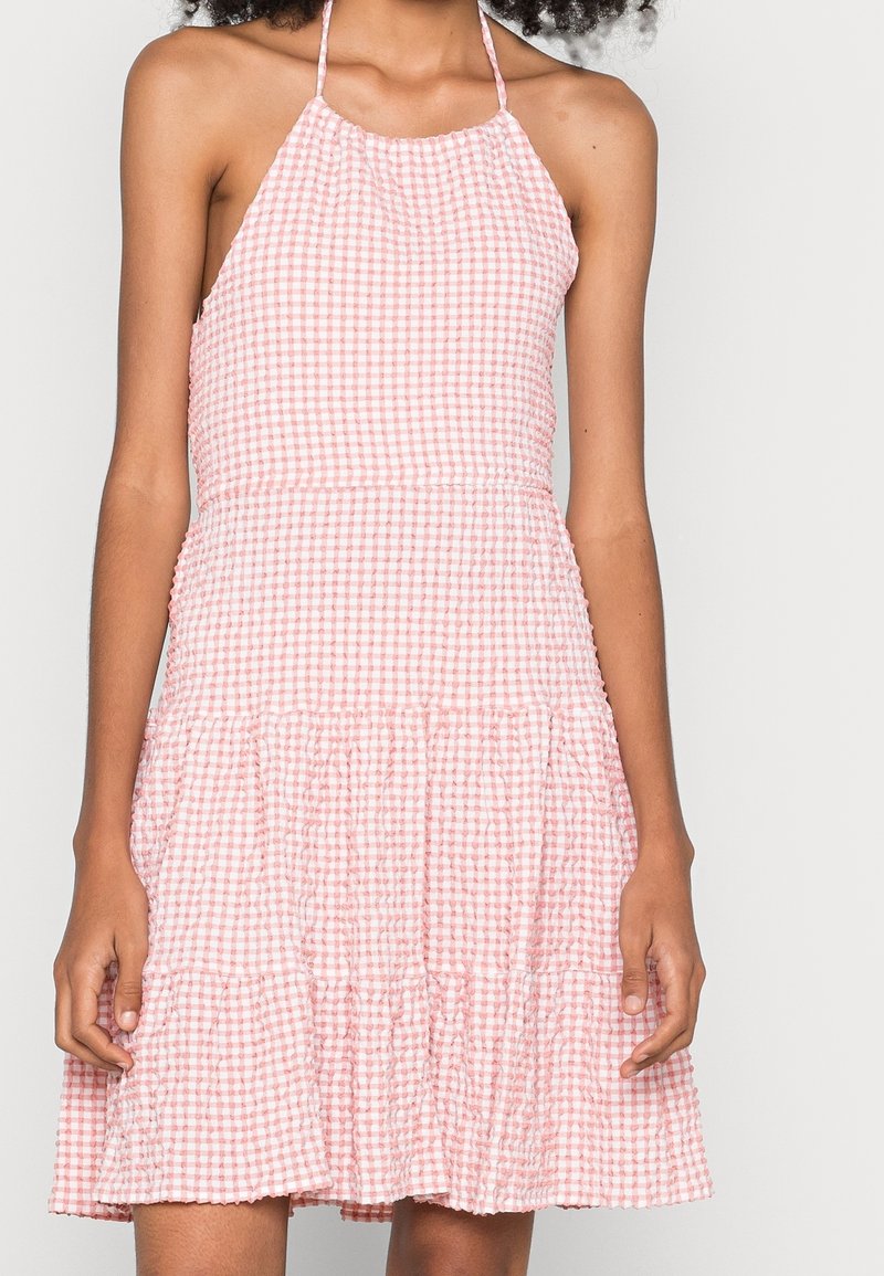 Femme portant une robe dos-nu vichy rose et blanche avec une jupe à volants, debout sur un fond clair uni.
