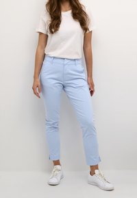 Femme portant un pantalon slim court bleu clair, un t-shirt blanc et des baskets blanches sur un fond clair uni.