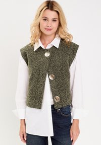 Un gilet en laine vert avec une finition texturée, présentant une ouverture à l'avant et trois grands boutons argentés brillants. Porté par-dessus une chemise blanche.