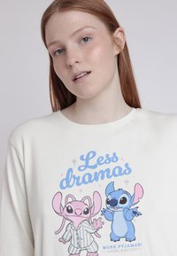 Sudadera blanca de gran tamaño hecha de algodón suave, con una impresión gráfica de personajes de dibujos animados en tonos rosa y azul. El texto dice "¡Menos dramas!"
