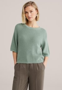 Pull en maille vert menthe doux, avec un col rond, des manches trois-quarts et un motif texturé, associé à un pantalon gris à jambes larges.