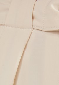 Světlá béžová látka s hladkou, matnou texturou, zdobená prošitým záhybem a širokým pasem. Detailní záběr materiálu a designu.