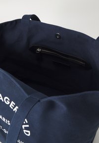 Saco de lona azul-marinho com um bolso com fecho no interior. Possui alças robustas e um fecho de pressão, com letras brancas de um lado.
