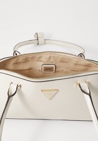 Cremefarvet håndtaske med en glat, blank finish. Udstyret med guldfarvet hardware, lynlåslukning og en beige indvendig, der har "GUESS" logo mønster.