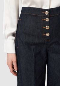 Tmavé džíny s vysokým pasem, ozdobené čtyřmi stříbrnými knoflíky a kontrastním oranžovým prošíváním. Měkká textura s přiléhavým střihem.