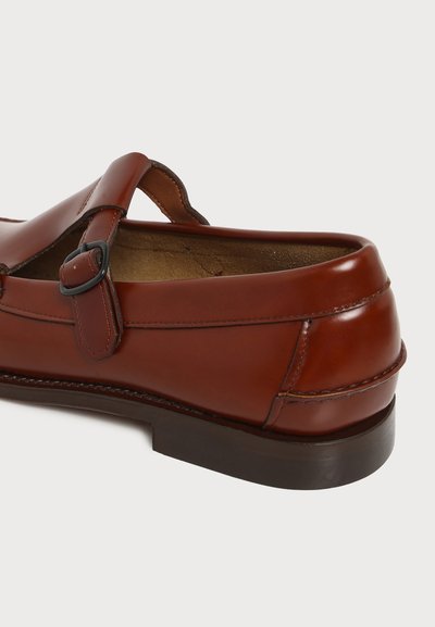 Chaussure mocassin en cuir marron avec une bride à boucle et une semelle marron foncé, vue de l’arrière sur un fond clair uni.
