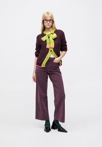 Cardigan din tricot în violet închis cu o fundă verde și accente roz, asortat cu pantaloni largi culoarea prunei și pantofi negri. Finisaj texturat.