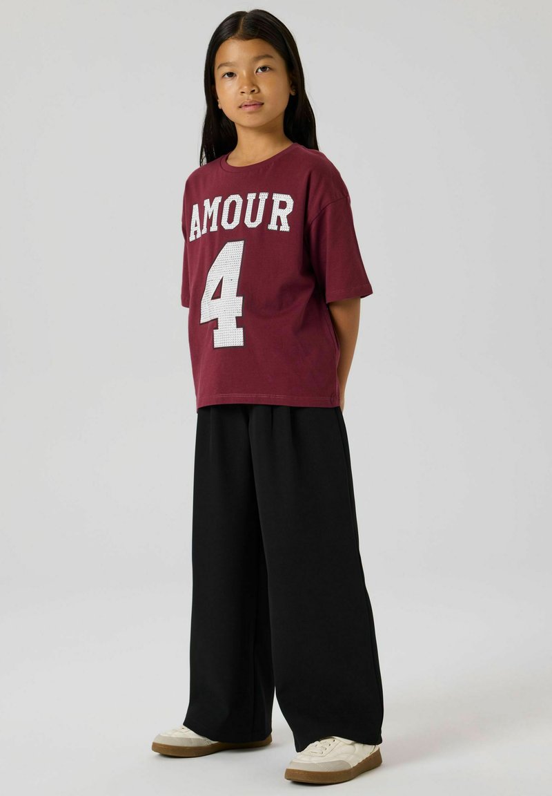 T-shirt en coton bordeaux avec le texte blanc "AMOUR" et le numéro "4" au dos, associé à un pantalon noir ample et des baskets crème.