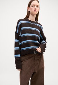 Braun gestreifter Pullover mit blauen horizontalen Linien, langen Ärmeln und lockerer Passform. Getragen mit braunen Cordhosen mit vertikalen Rippungen.