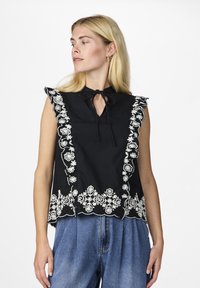 Svart ermeløs bluse med høy hals og knytting, med hvit blomstermønsterbroderi langs kantene og kanten. Bølgeformet trim fremhever.