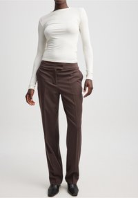 Langärmeliges weißes eng anliegendes Oberteil, kombiniert mit braunen maßgeschneiderten Hosen, die über seitliche Taschen und eine glatte Textur verfügen; schwarze High Heels.