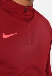 Haut Nike à quart de fermeture éclair en tissu bordeaux, avec un logo rose. Texture lisse avec un col montant et un détail de petite fermeture éclair.