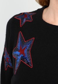 Schwarzer Pullover mit mehrfarbigen Stern-Applikationen aus roten und blauen Pailletten. Hat einen runden Halsausschnitt und eine weiche Textur.