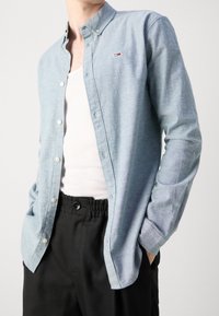 Mies, jolla on päällään vaaleansininen napillinen paita, valkoinen toppi ja mustat housut, kädet taskuissa, yksinkertaista valkoista taustaa vasten.