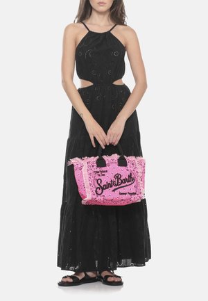 Donna che indossa un abito lungo nero con ritagli e sandali neri, che tiene una borsa rosa con frange con la scritta "Saint Barth".