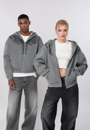 Du modeliai dėvi pilkus džemperius su gaubtu, ant pečių – juodos sparnų raštai; vienas derina džemperį su baltu marškinėliu ir pilkomis džinsinėmis kelnėmis, kitas – su balta trumpa palaidine ir juodomis džinsinėmis kelnėmis.