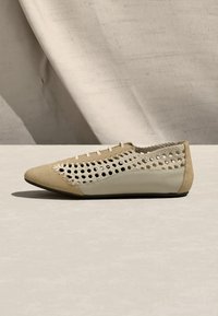 Chaussure plate beige avec tige en maille ouverte tissée, bout pointu et semelle solide sur une surface neutre claire avec un arrière-plan en tissu.