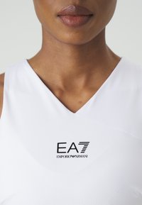 Bílé sportovní tílko bez rukávů s V výstřihem. Na přední straně se nachází černé logo EA7 Emporio Armani, hladká tkanina a přiléhavý střih.