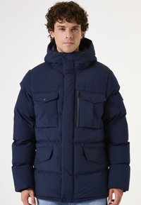 Marineblauwe gewatteerde jas met een hoge kraag, meerdere voorzakken en een afneembare capuchon voor warmte en functionaliteit.