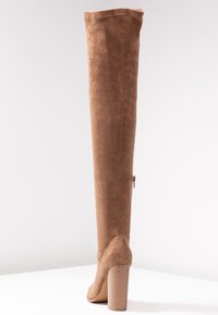Bota de ante color tostado, sobre la rodilla, con un tacón grueso acanalado y cremallera lateral. Textura suave, con un diseño ajustado y punta redonda.